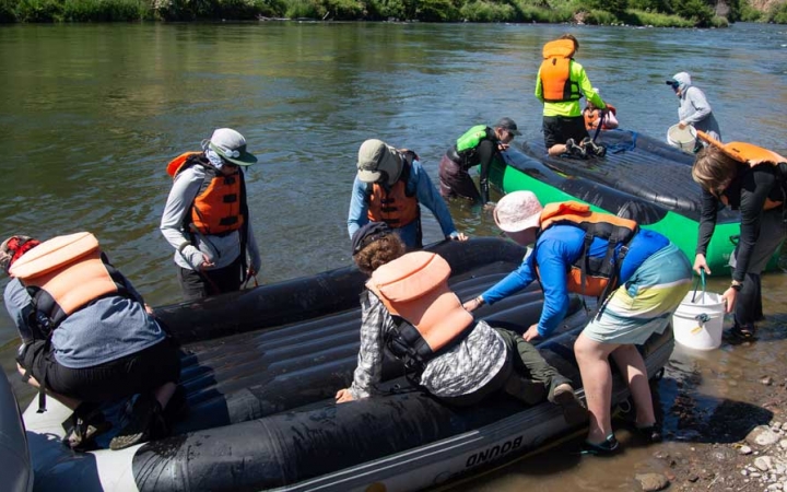 A group of people wearing life jackets appear to clean the bottom of rafts in ankle-deep water.